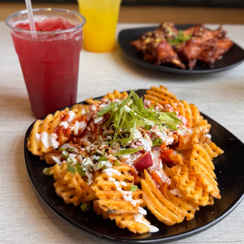 Waffle fries loaded with sauces and green onions, with drinks and chicken wings in the background.