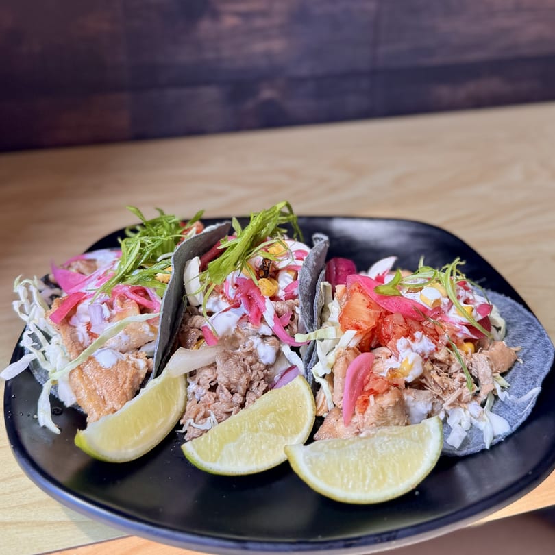 Three Korean-fusion tacos with various fillings, sauces, and lime wedges on a black plate.