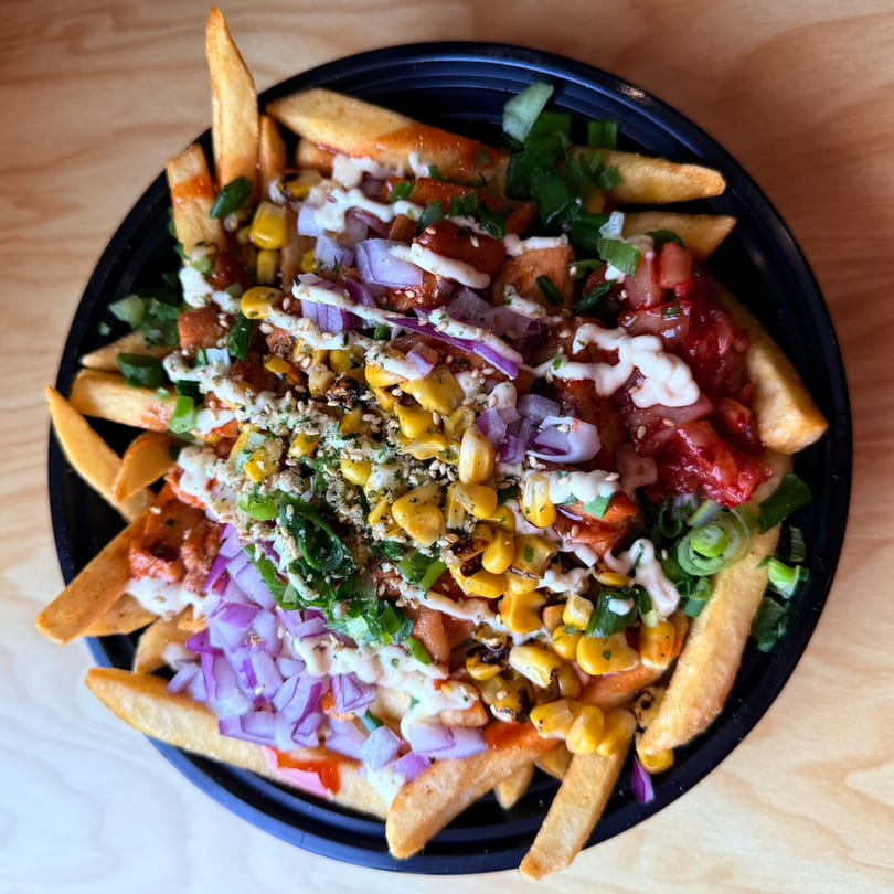A black bowl of Korean-fusion fries topped with kimchi, corn, red onions, green onions, and sauces.
