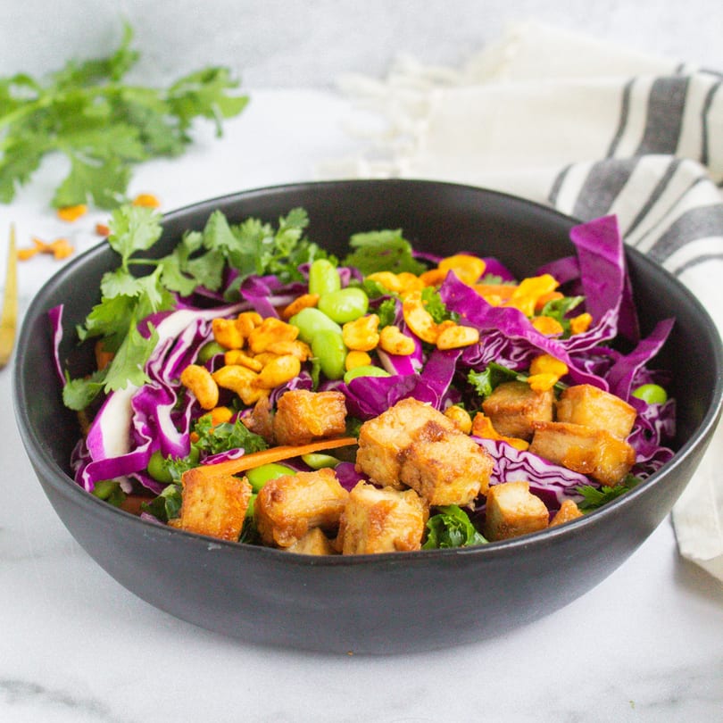 A vibrant Korean-fusion salad with crispy tofu, purple cabbage, edamame, and cashews in a black bowl.
